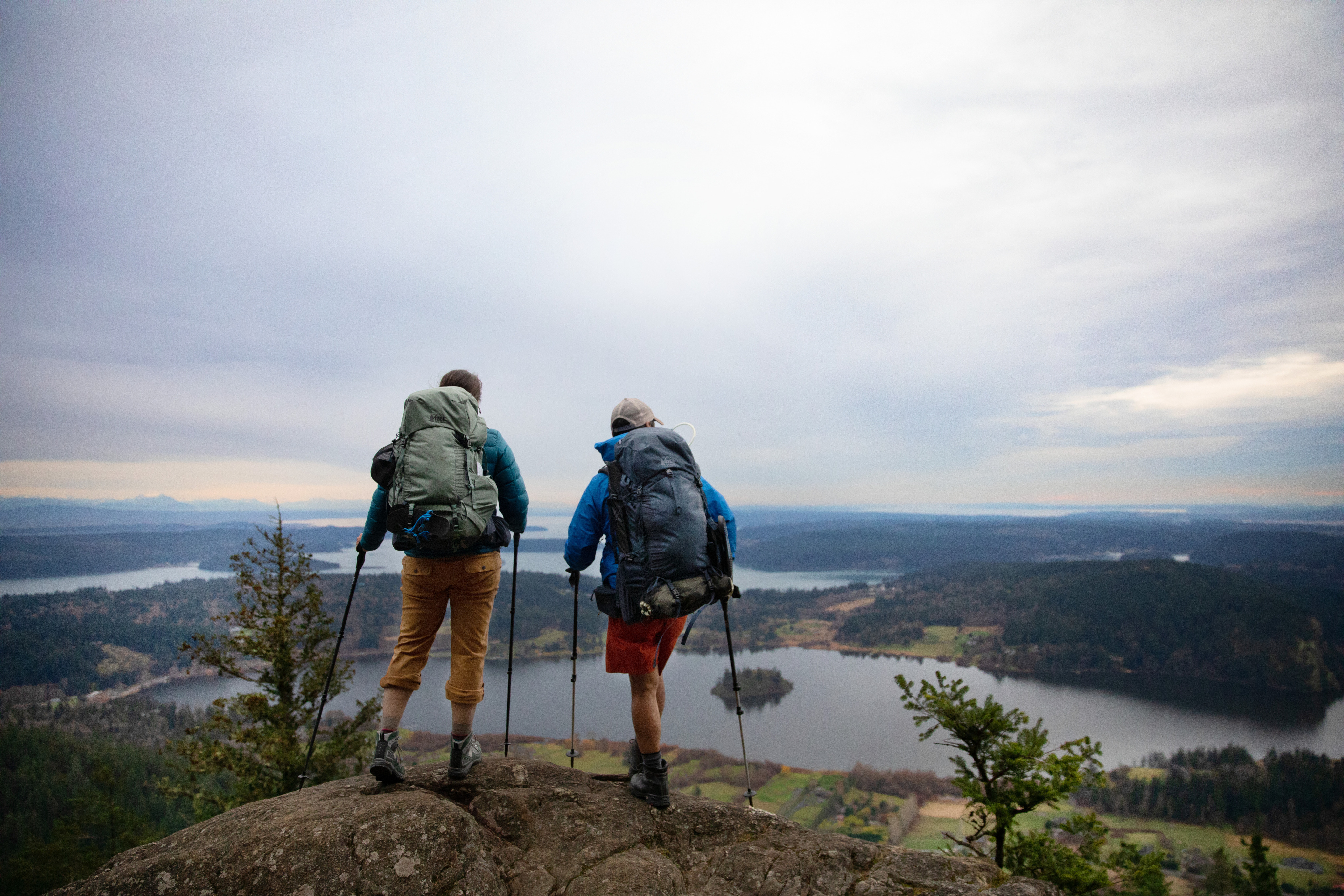 Two people lean on their trekking poles, taking a break from backpacking, and look out at the view of the water below them.