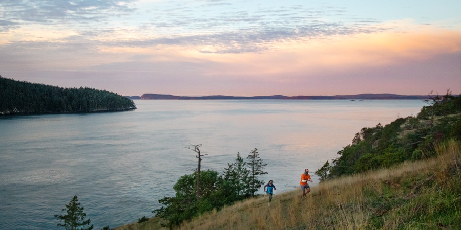 Two runners make their way along a ridge line above a sound at sunset.