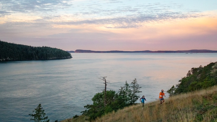 Two runners make their way along a ridge line above a sound at sunset.