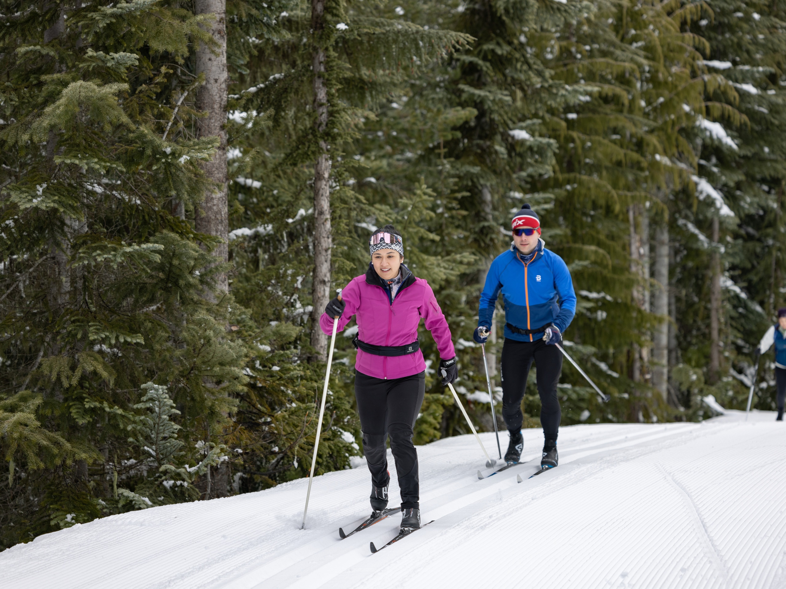 Two people cross country ski along a groomed trail.