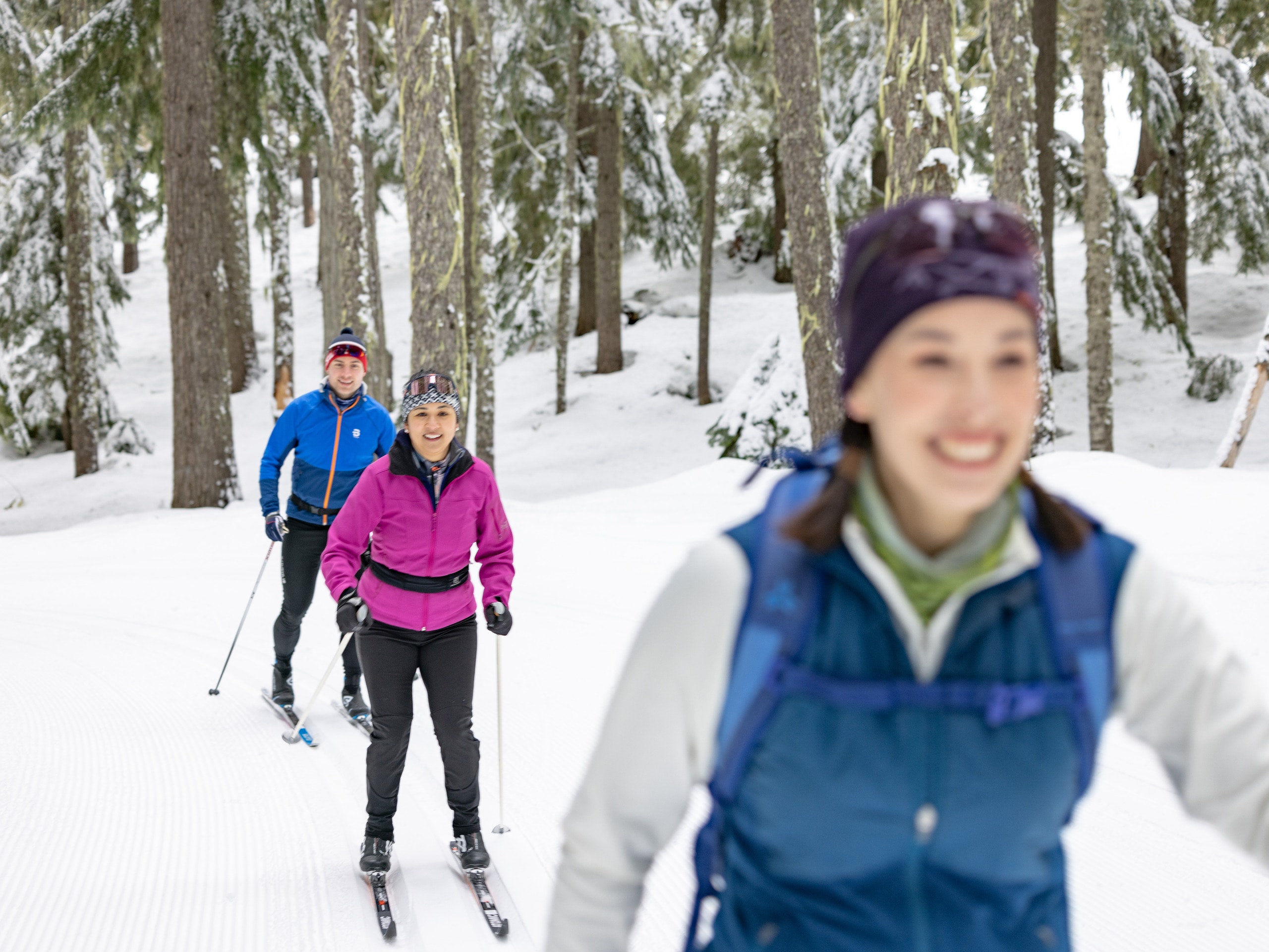 Three smiling people cross-country ski in a wooded area.