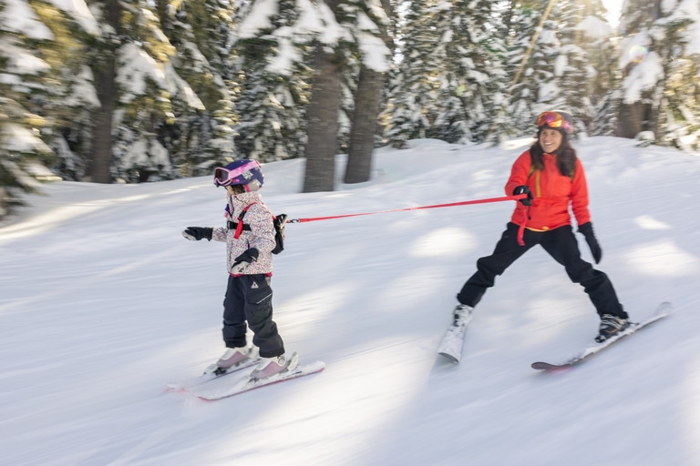 A parent helps a child ski using a ski leash. Kids' downhill skis fit the youngest rippers.