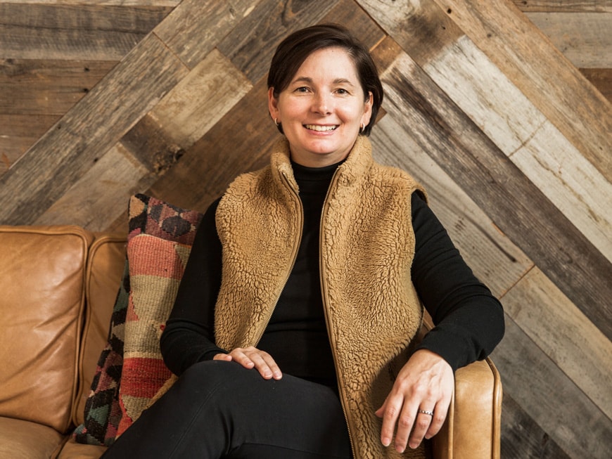 Michelle Cutter, seated, wearing a brown fleece vest over a black shirt. She sits on a leather couch in front of a patchwork wood wall.