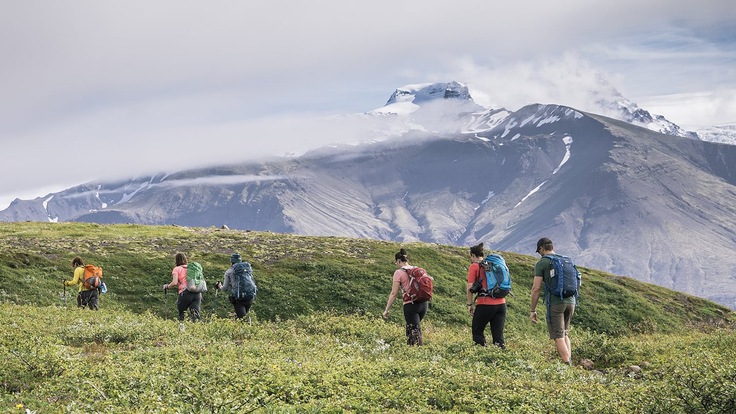 A group of hikers on a trail atop a green knoll with snowcapped peaks in the distance.