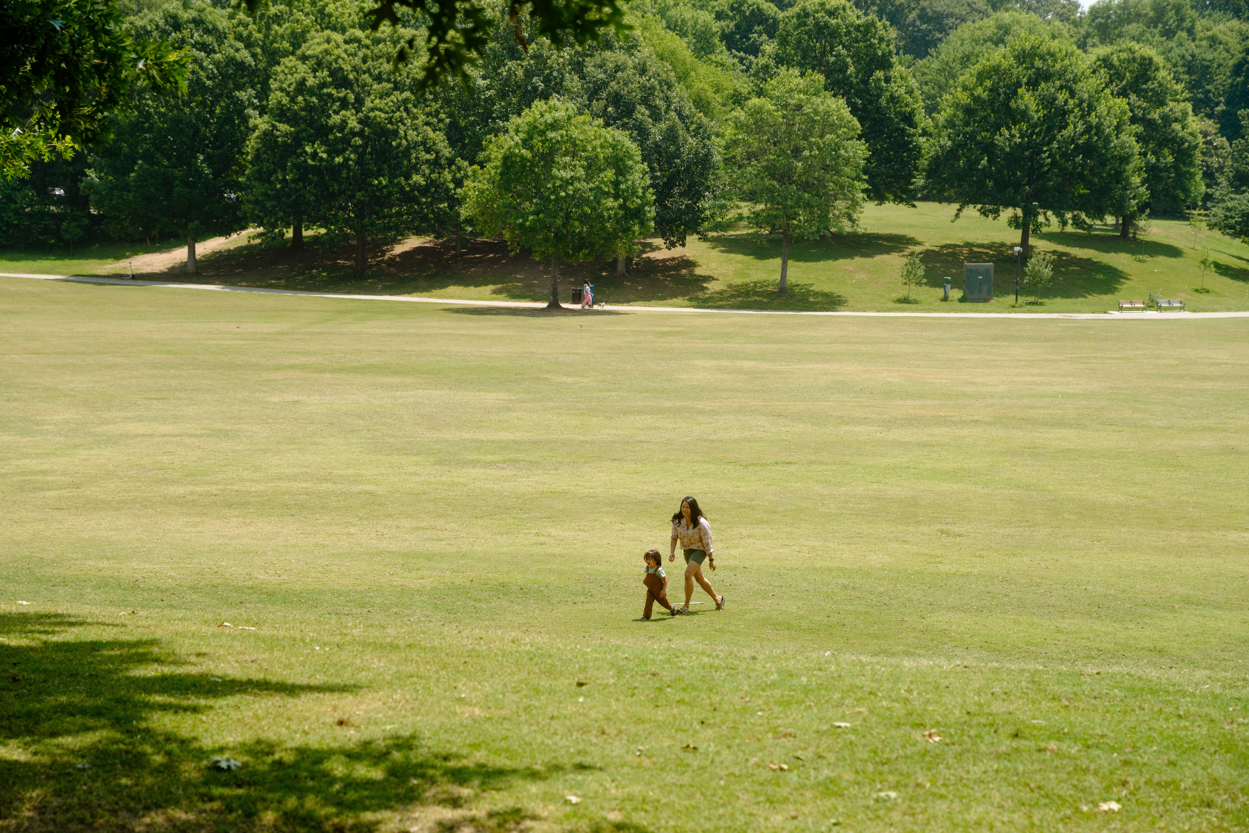 A woman walks with a child across a large, grassy park. Trees line the background under a clear sky.