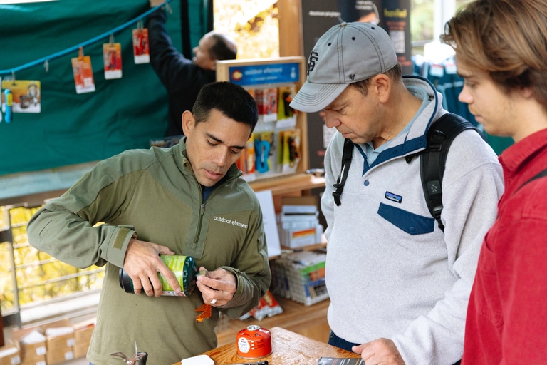 A founder demonstrates how to use a tool to open a can of propane to two customers in an open-air marketplace