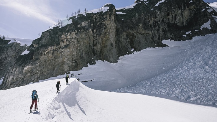 wide shot of 3 skiers trekking up a mountain on a sunny day