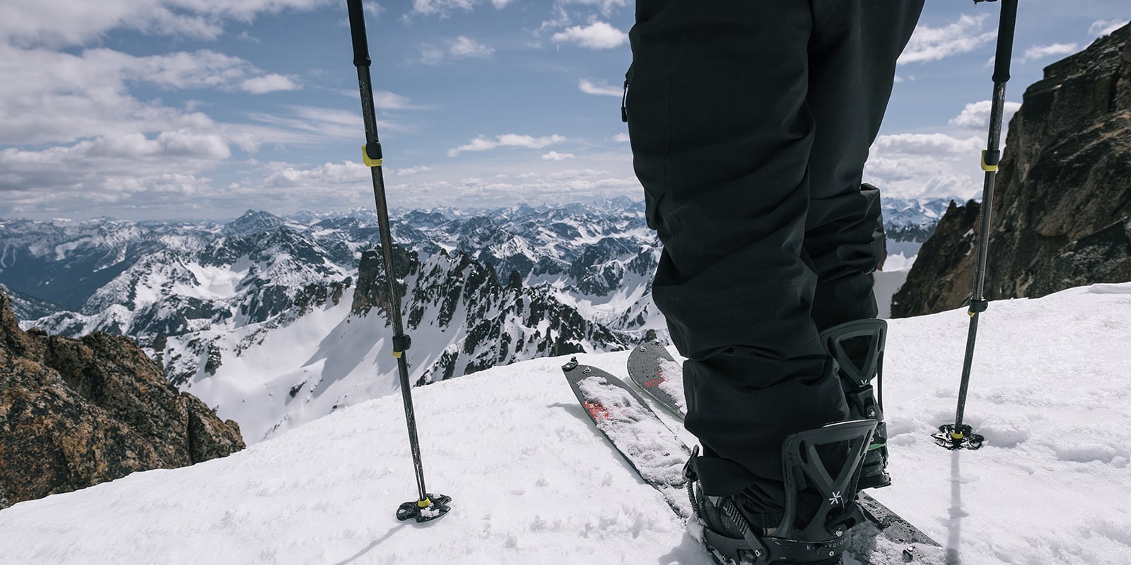  Close up of a skier's lower legs, boots, ski's and ski poles at the top of a ski run overlooking a beautiful mountain range and partially cloudy sky