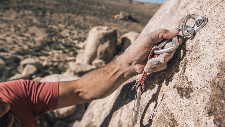 Man wearing a helmet clipping a carabiner onto a climbing bolt that's attached to a rock face.