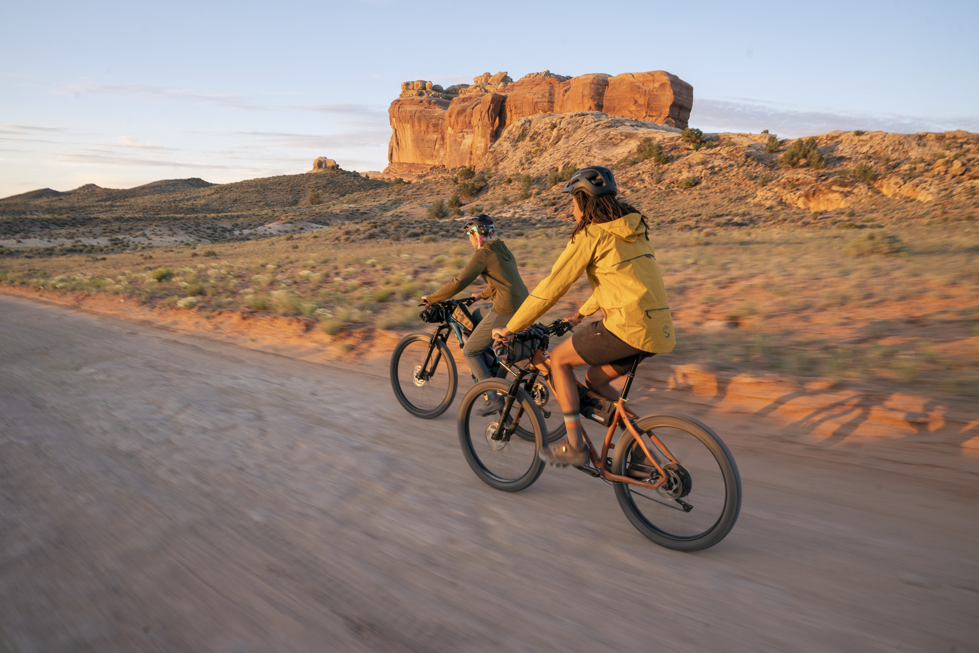 Two people bike along a desert trail with canyons in the background.