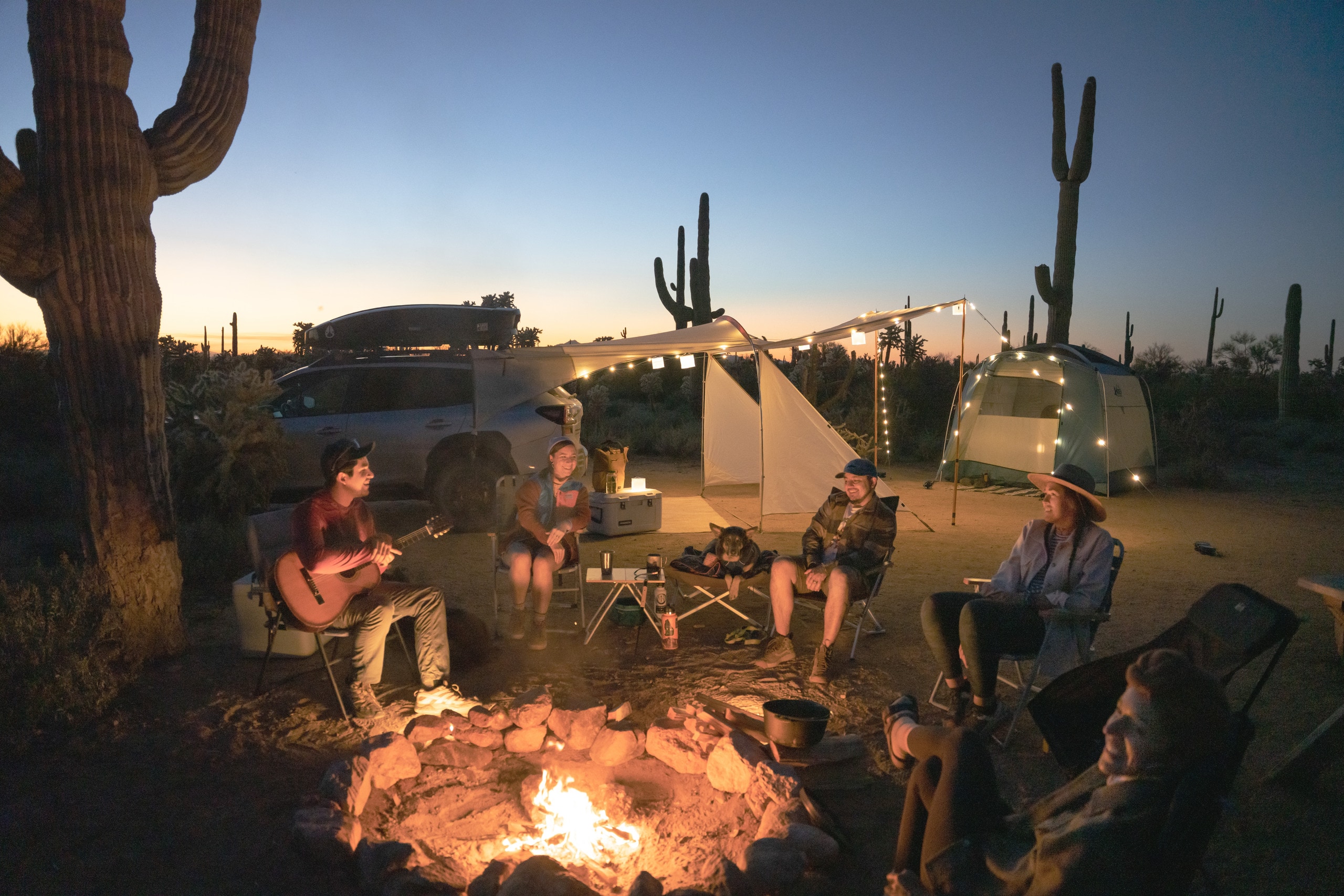 Picture of a campsite, featuring a tent, sun shade and campers gathered around a campfire