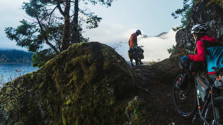 A man and woman walk their bikes up a single track trail while bikepacking