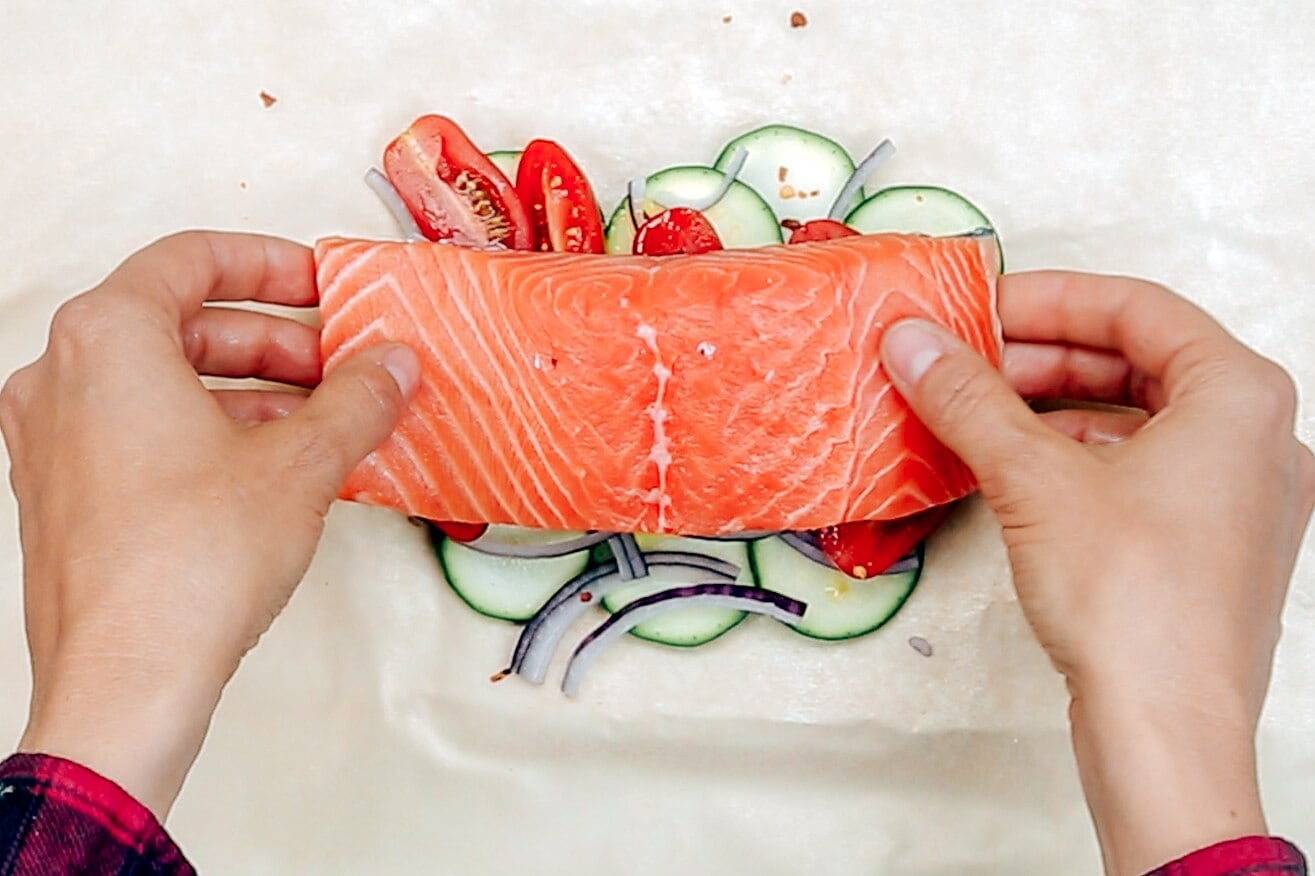 Two hands placing a salmon filet on top of cherry tomatoes and zucchini.