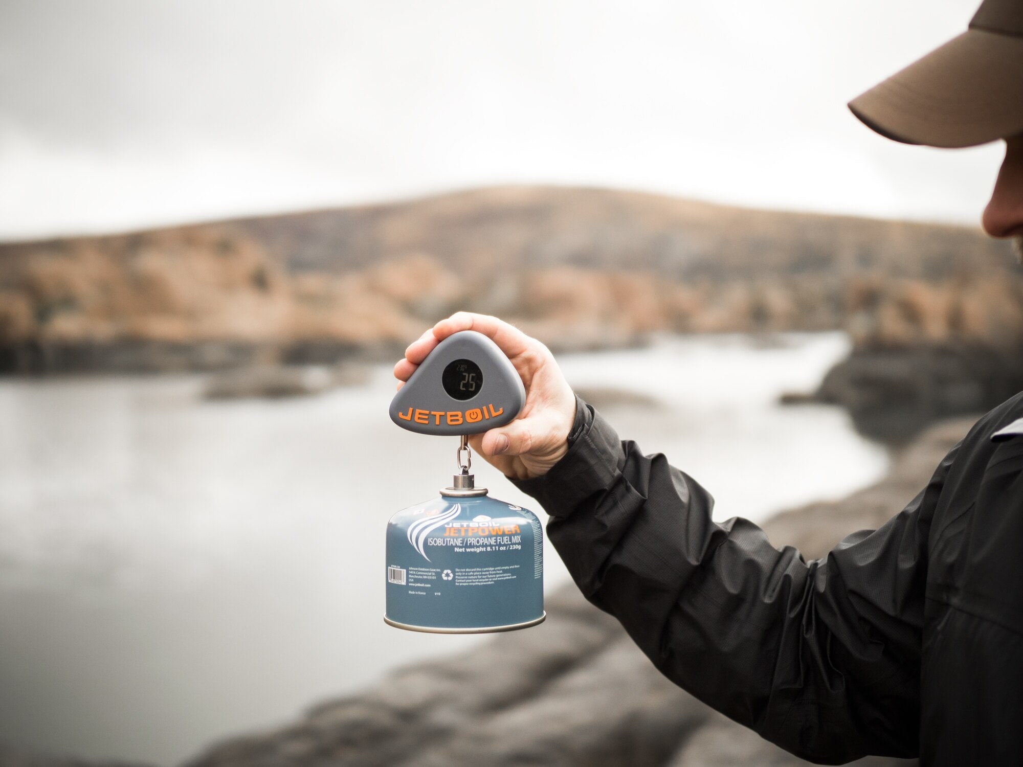 Person holds scale with a fuel canister dangling in front of a peaceful river.