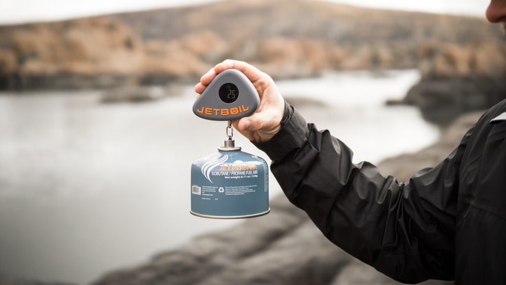 Person holds scale with a fuel canister dangling in front of a peaceful river.