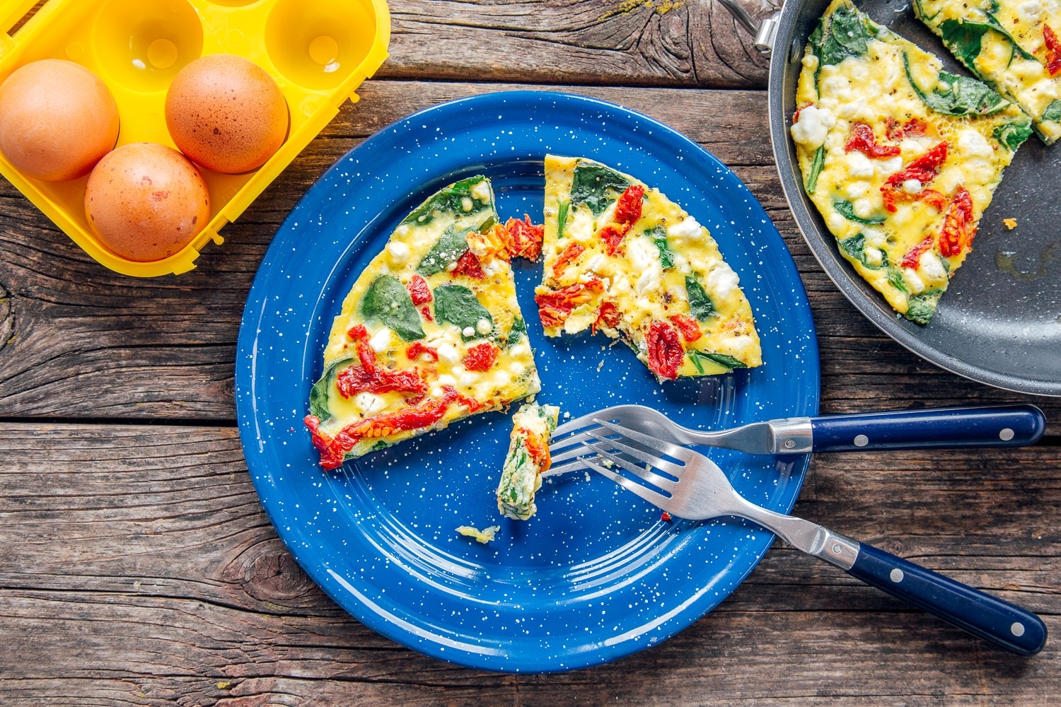 Two colorful slices of frittata sit on a blue-speckled camp plate along with two forks and a plastic carton of eggs.