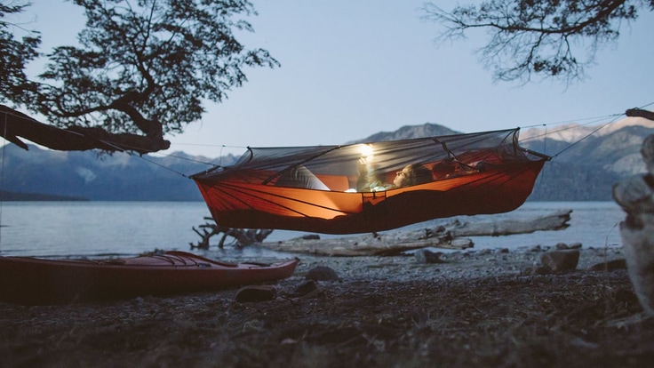 A camper in a hammock tent reads by a light hung at the top of the net covering.