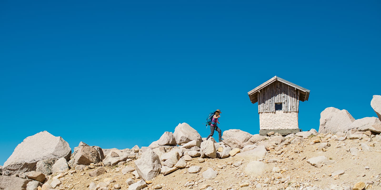 A person with a backpack traverses some rocks leading toward a small hut.