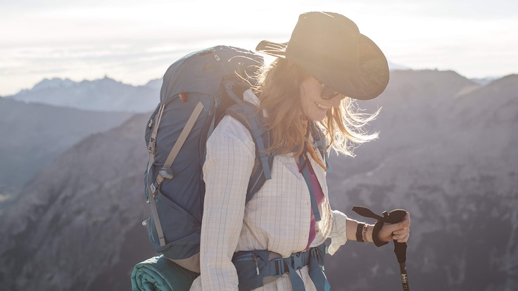 A long-haired hiker who is backlit by sunlight and wearing a pack, wide-brim sun hat and long-sleeve sun shirt