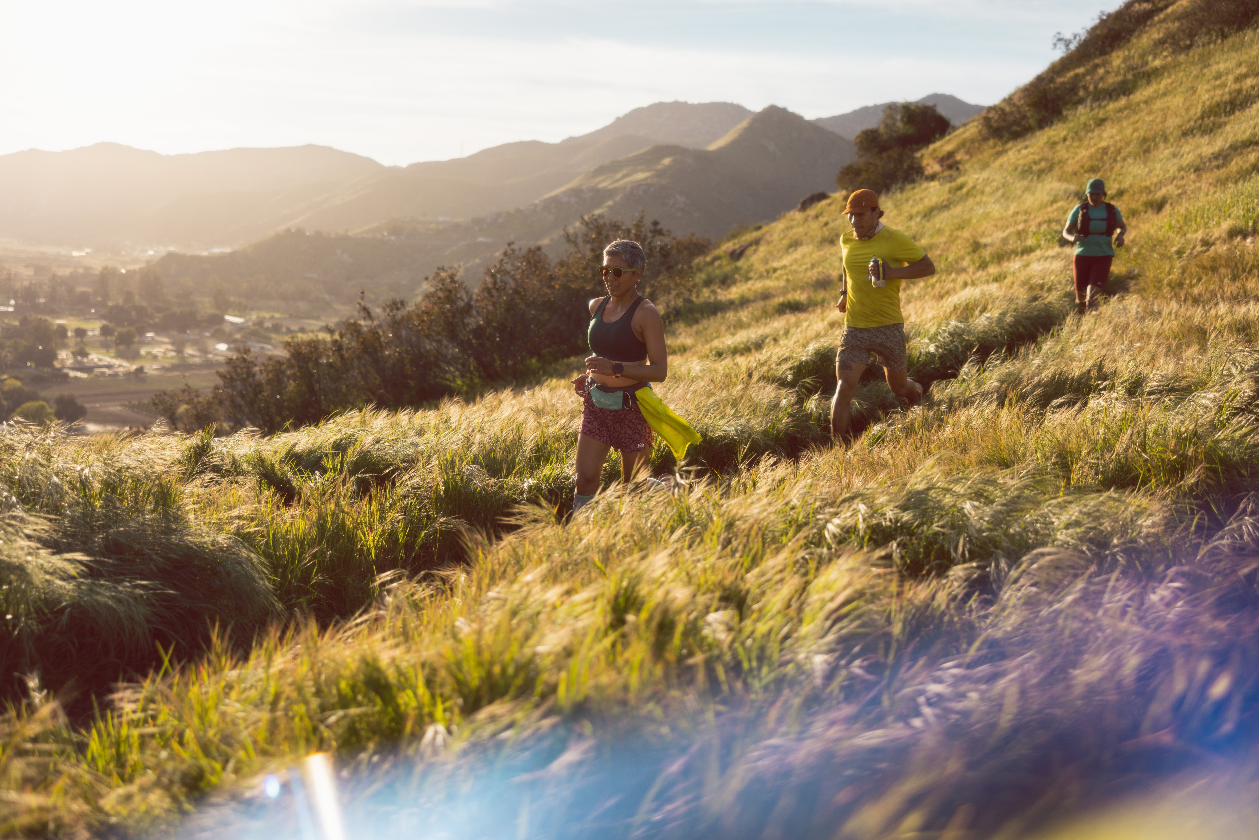 Three people run through a trail on a grassy hillside.