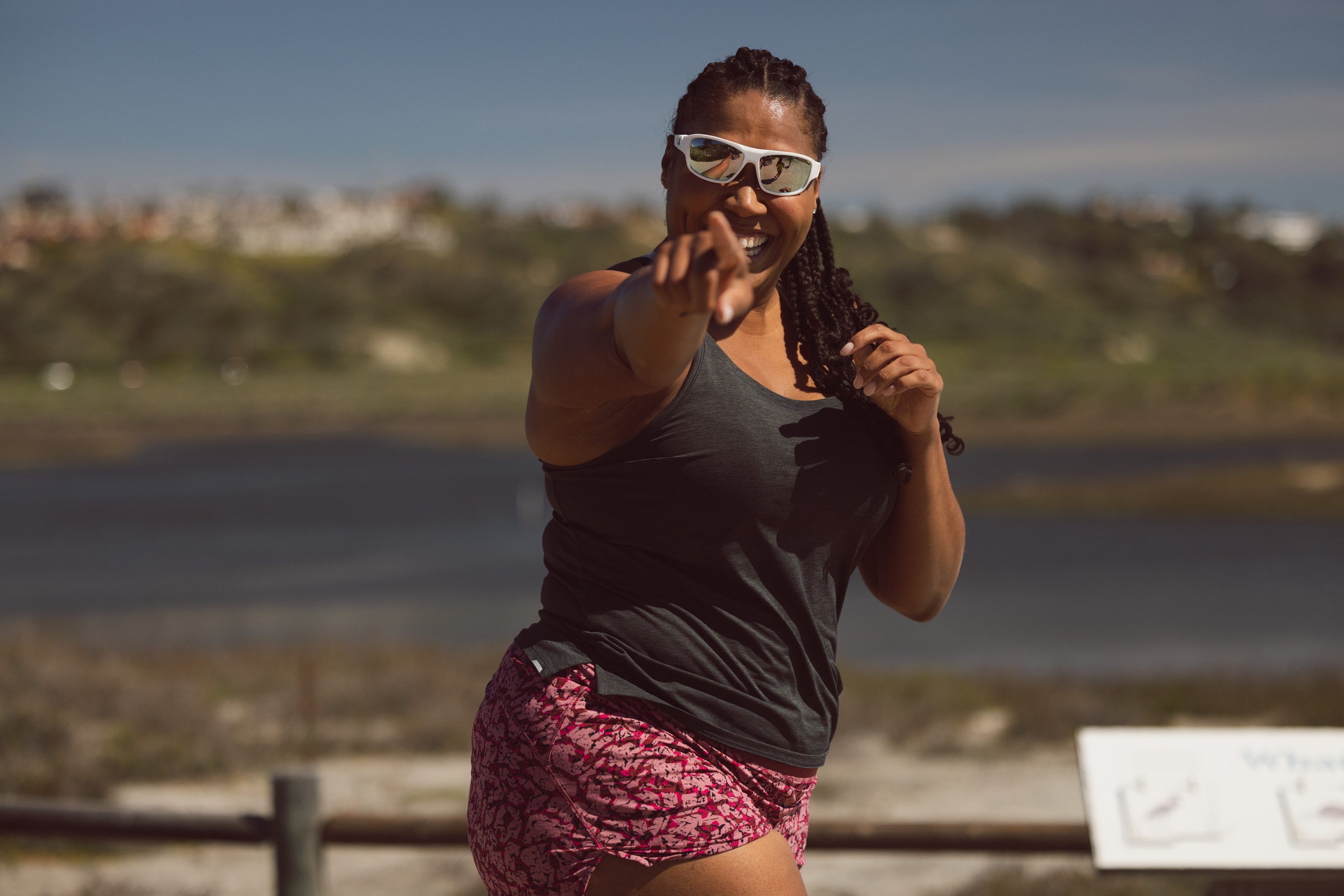 A person in pink patterned running shorts and a charcoal black running tank top and white sunglasses runs while pointing to the camera and smiling