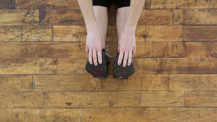A woman seated on a wooden floor reaches to touch her toes
