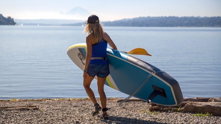 A woman carries her stand up paddle board towards water