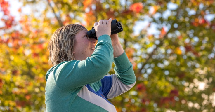 A woman looking through binoculars with fall foliage in the background