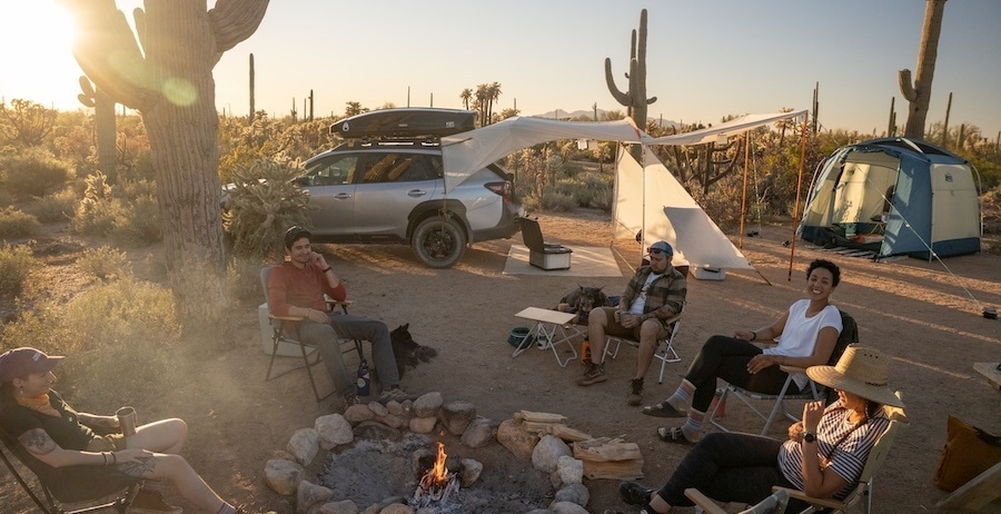 Campers sit around a fire in a desert setting. In the background is a car with a vehicle shelter attached and a tent.