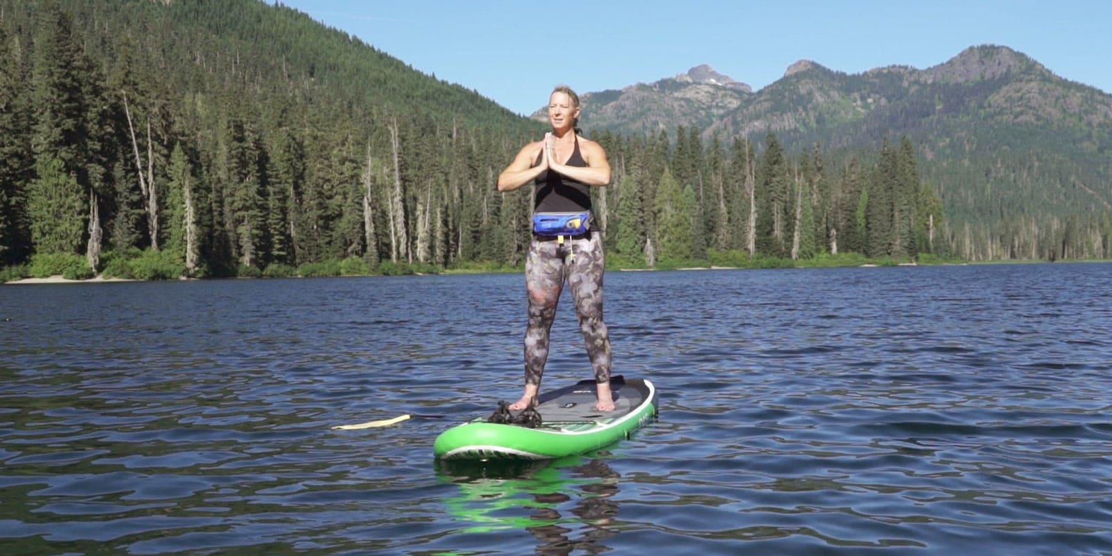 A person doing yoga stands in Mountain Pose on a stand up paddle board on a lake.